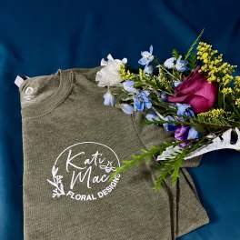 Folded green logo t-shirt with a small bouquet of pink rose and blue and yellow flowers on top