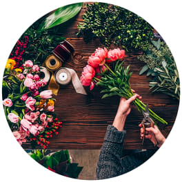 Hands trimming pink flowers beside ribbons and greenery on a wooden table