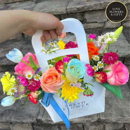 Colorful mixed bouquet of roses, daisies, and mums in a white carry bag with handle