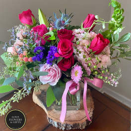 Pink and purple mixed flower arrangement with roses and hydrangea in a glass vase on a wood slice stand.