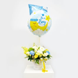 Blue baby boy balloon above a white basket of yellow and white flowers