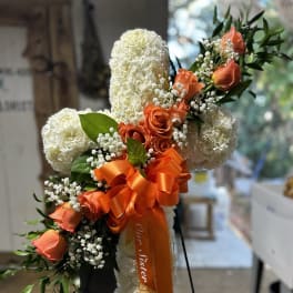Standing white floral cross with orange roses, ribbon, and greenery on a metal easel.