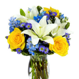 Mixed bouquet of white lilies, yellow roses, and blue flowers arranged in a clear glass vase