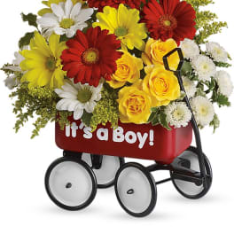 Mixed bouquet of red, yellow, and white flowers arranged in a red wagon that reads It’s a Boy.