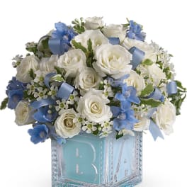 Compact arrangement of white roses and blue flowers in a blue baby block glass cube vase.