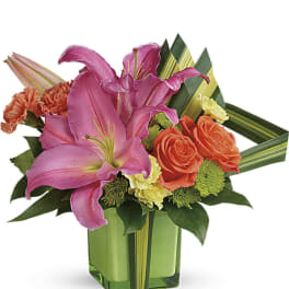 Bright arrangement of pink lilies, orange roses and carnations in a green cube vase with striped leaves.