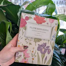 Hand holding a floral-patterned book in front of large green leaves