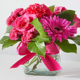 Pink roses, carnations, and a gerbera daisy in a glass vase with a pink ribbon