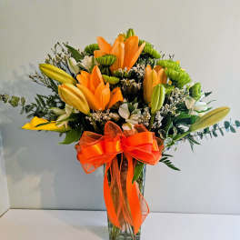 Orange lilies and green chrysanthemums in a clear glass vase with an orange ribbon