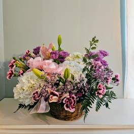 Basket arrangement of white hydrangeas, pink lilies, and purple flowers with a pink ribbon