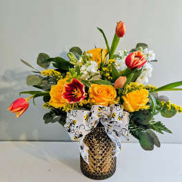 Yellow roses and tulips arranged in a gold vase with a bee-patterned ribbon