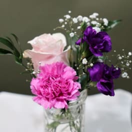 Small arrangement with a pink rose, bright pink carnation, and purple blooms in a clear glass jar.