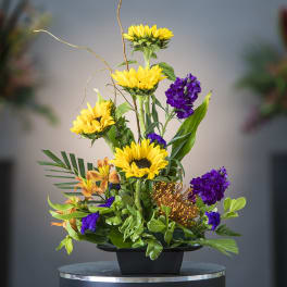 Tall floral arrangement with yellow sunflowers and purple blooms in a black container