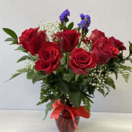 Red roses with white and purple filler flowers in a red vase with a red ribbon bow