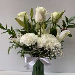 Tall arrangement of white roses, lilies, and carnations in a green glass vase with a white bow