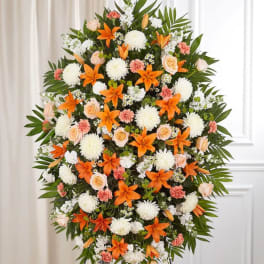 Tall standing spray with orange lilies, white mums, peach roses, and carnations on a leafy easel.