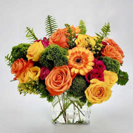 Bright arrangement of orange and yellow roses with gerbera daisies in a clear glass cube vase