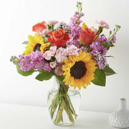 Mixed bouquet of sunflowers, orange roses, and pink blooms arranged in a clear glass vase.