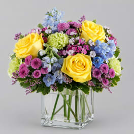 Mixed bouquet of yellow roses, blue flowers, and purple blooms in a clear glass vase