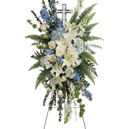 Standing floral spray with white lilies, cream roses, and blue flowers around a cross
