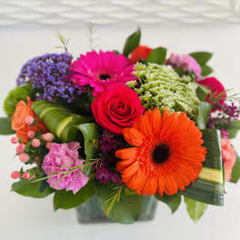 Bright mixed bouquet of orange gerbera daisies, hot pink roses and other blooms in a low glass cube vase