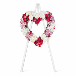 Heart-shaped floral wreath on a white easel with red, pink, and white flowers.