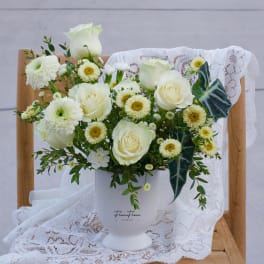 White roses and daisy-like flowers arranged in a white vase with lace draping.