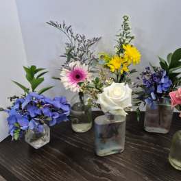 Five small floral arrangements in glass vases with blue, pink, white, and yellow flowers