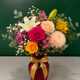Mixed bouquet of roses, lilies, and daisies in a red vase with a gold ribbon
