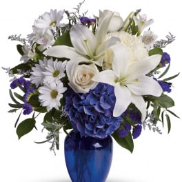 White lilies, roses, and daisies with blue hydrangea in a deep blue glass vase
