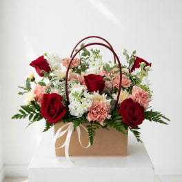 Basket arrangement of red roses, pink carnations, and white flowers with ribbon handles
