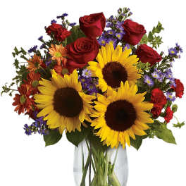 Clear glass vase of yellow sunflowers with red roses, red carnations, and purple and orange accent flowers