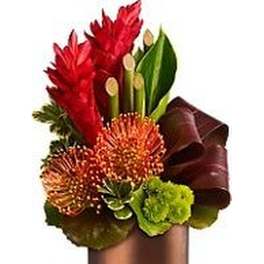 Tropical arrangement with red and orange flowers, green accents, and curled leaves in a bronze cylinder vase