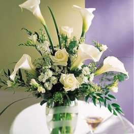 Tall arrangement of white calla lilies and roses in a clear glass vase with green ribbon accents