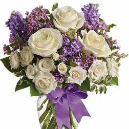 Arrangement of white roses and purple flowers in a clear glass vase with a purple ribbon.
