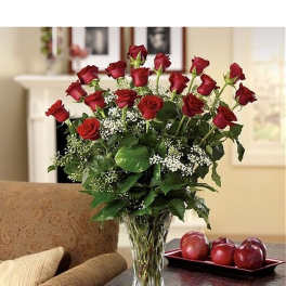 Tall arrangement of long-stem red roses with small white filler flowers in a clear glass vase