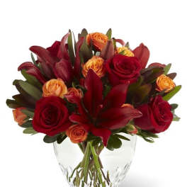 Compact arrangement of red lilies and red and orange roses in a clear glass vase