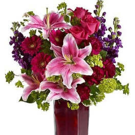 Tall arrangement of pink lilies, red roses, purple blooms and green accents in a clear glass vase