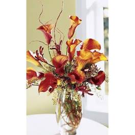Tall arrangement of orange and red calla lilies in a clear glass vase with curly branches.