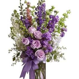 Tall arrangement of lavender roses and purple and white flowers in a clear glass vase with a purple bow