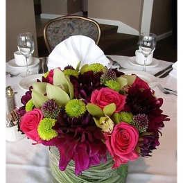 Low centerpiece of pink roses, dark purple blooms, green mums and orchids in a glass cylinder vase