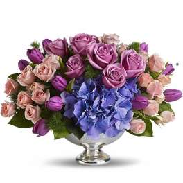 Low centerpiece of purple roses, pink spray roses, tulips, and blue hydrangeas in a silver pedestal bowl