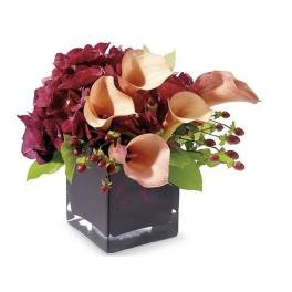 Low arrangement of peach calla lilies and burgundy flowers in a dark square glass vase