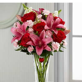 Tall clear vase of red roses, pink lilies, and light pink carnations in a mixed bouquet