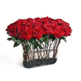 Rectangular vase filled with many red roses arranged in tight rows over black stones.