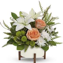 Low arrangement of peach roses, white lilies, and green mums in a white bowl on a wooden stand