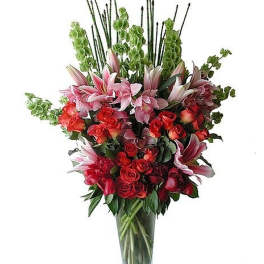 Tall arrangement of pink lilies, red roses, and green bells of Ireland in a clear glass vase.