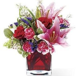 Pink lilies, red roses, and pink carnations arranged in a red glass cube vase