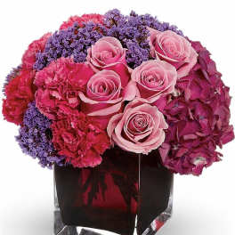 Compact arrangement of pink roses, carnations and purple flowers in a clear glass cube vase