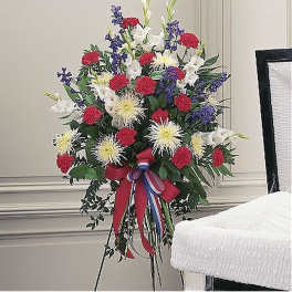 Standing red, white, and blue funeral spray on an easel with bow beside a white casket.